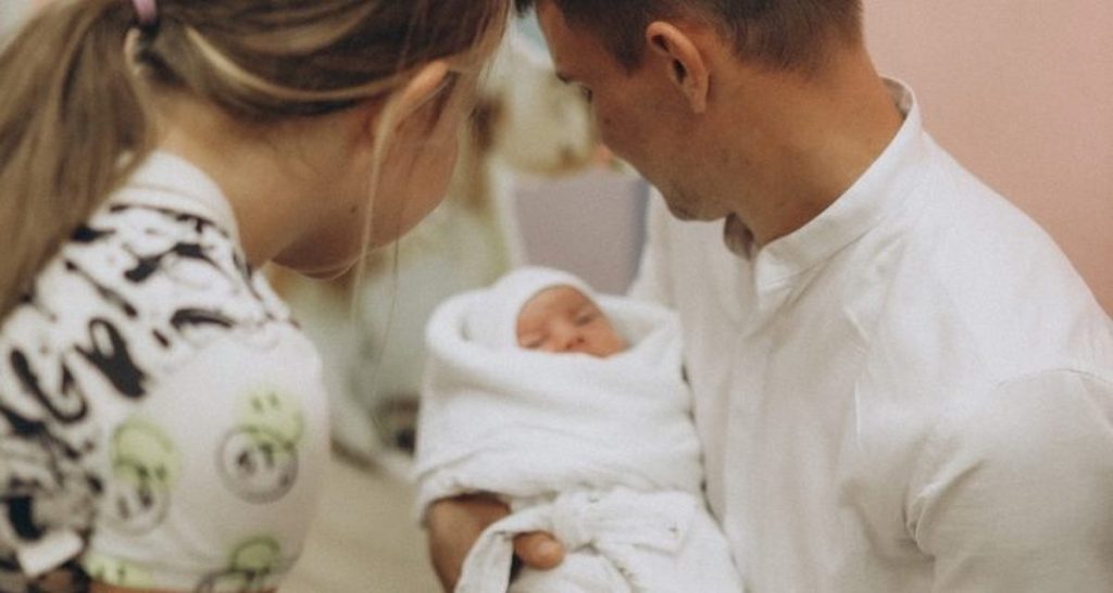 Мужчина держит младенца на руках, женщина склонилась рядом – семья после роддома.